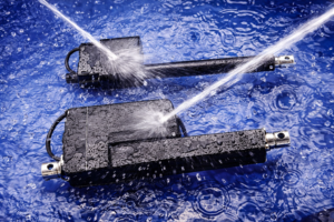 Energie besparen in foodmachines: waarom steeds meer OEM’s afscheid nemen van pneumatiek | Axis & Stuifmeel Twee elektrische lineaire actuatoren worden onder hoge druk gereinigd met waterstralen op een natte blauwe ondergrond, waarbij waterdruppels en robuuste afdichting de geschiktheid voor hygiënische omgevingen benadrukken.